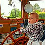 baby, blue_eyes, boat, building, child, controls, curious, dashboard, hand, holding, hoodie, indoor, instrument_panel, leopard_print, nature, person, steering_wheel, trees, window, wood
