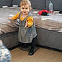 toddler, child, standing, dress, plaid, yellow_shirt, black_shoes, couch, living_room, baby_bottle, clothing, floor, smiling, person, indoors, furniture, cute, young_child, home, playful