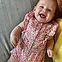 baby, smiling, infant, floral_clothing, romper, couch, pillow, happy, lying_down, indoors, cute, child, person, skin, face, cheerful, baby_girl, cozy, comfort, bright_eyes