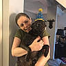 animal, black_dog, casual_clothing, celebration, dog, friendship, furniture, happy, headwear, home_interior, hugging, indoor, lamp, living_room, party_hat, person, pet, smiling, wall, woman