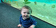 Bennett is registered to the contest to win money with this photo: toddler, child, aquarium, fish, blue_striped_shirt, gray_pants, black_socks, indoor, floor, sitting, smiling, curious, aquatic_plants, tank, water, yellow_fish, young_child, happy, portrait, playful