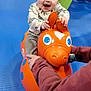 Bennett is registered to the contest to win money with this photo: toddler, child, toy, orange, bouncy_horse, indoor, play, smile, happy, person, adult_hand, floor, blue_floor, clothing, long_sleeve, pants, playground, fun, support, expression