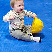 Bennett is registered to the contest to win money with this photo: toddler, child, indoor, blue_floor, hard_hat, yellow, dinosaur_print, long_sleeve_shirt, gray_pants, socks, sitting, happy, smiling, playful, young_child, floor_mat, toy, casual_clothing, person, cute