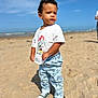beach, casual_clothing, child, confident, curly_hair, daytime, expression, footwear, mickey_mouse, outdoor, pants, person, sand, sky, sneakers, standing, sunny, toddler, tshirt, vacation
