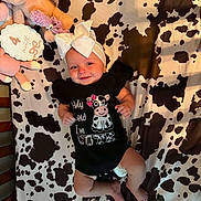 Brinley is registered to the contest to win money with this photo: baby, bed, black_clothing, bow, child, cow_pattern, crib, cute, fabric, headband, infant, laying, milestone, onesie, pink, plush, smile, soft_toy, toy, white_clothing