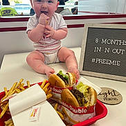 Eliana joined the competition — help win amazing prizes! baby, infant, bow, headband, table, fast_food, burger, fries, food, tray, sign, text, indoor, restaurant, happy, smiling, person, clothing, sitting, cute