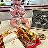 baby, infant, bow, headband, table, fast_food, burger, fries, food, tray, sign, text, indoor, restaurant, happy, smiling, person, clothing, sitting, cute