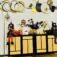 Left Eye is registered to the contest to win money with this photo: cat, cabinet, witch_doll, halloween_decorations, pumpkin, dried_flowers, vase, golden_mask, black_and_white_cat, decorative_cabinet, indoor, floor_lamp, orange_striped_tights, figurine, black_fur, purple_clothing, black_boots, wall_art, cozy, playful_cat