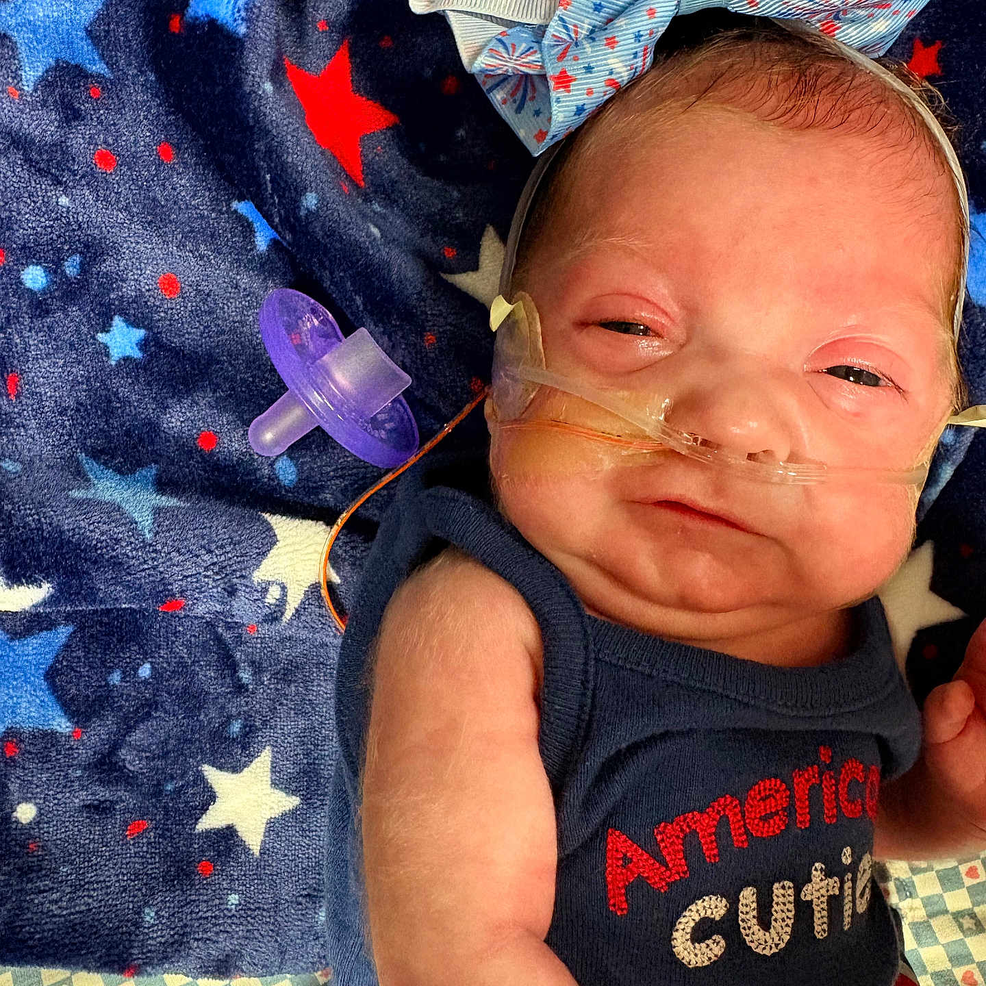 Eloise is registered to the contest to win money with this photo: arm, baby, blanket, blue, bow, clothing, cute, face, headband, infant, lying_down, newborn, oxygen_tube, pacifier, pattern, portrait, red, sleepy, stars, white