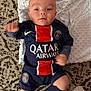 baby, blanket, blue_clothing, carpet, child, cute, football_kit, indoor, infant, looking_at_camera, lying_down, nike_logo, paris_saint_germain, patterned_carpet, person, small_fists, soccer_uniform, sock, soft_texture, sportswear