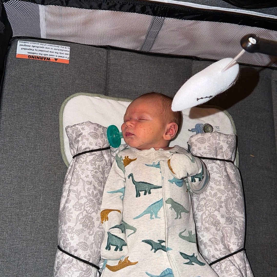 Samuel is registered to the contest to win money with this photo: baby, newborn, sleeping, onesie, dinosaur_pattern, playpen, blanket, pillow, mobile_toy, wooden_wall, cozy, infant, child, bedtime, nursery, soft_toy, resting, indoors, peaceful, cute