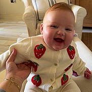 Matilda is registered to the contest to win money with this photo: baby, child, smiling, laughing, person, indoor, couch, armchair, hand, tattoo, bracelet, knitwear, strawberry_pattern, cozy, home, furniture, pillow, plant, wooden_furniture, floor