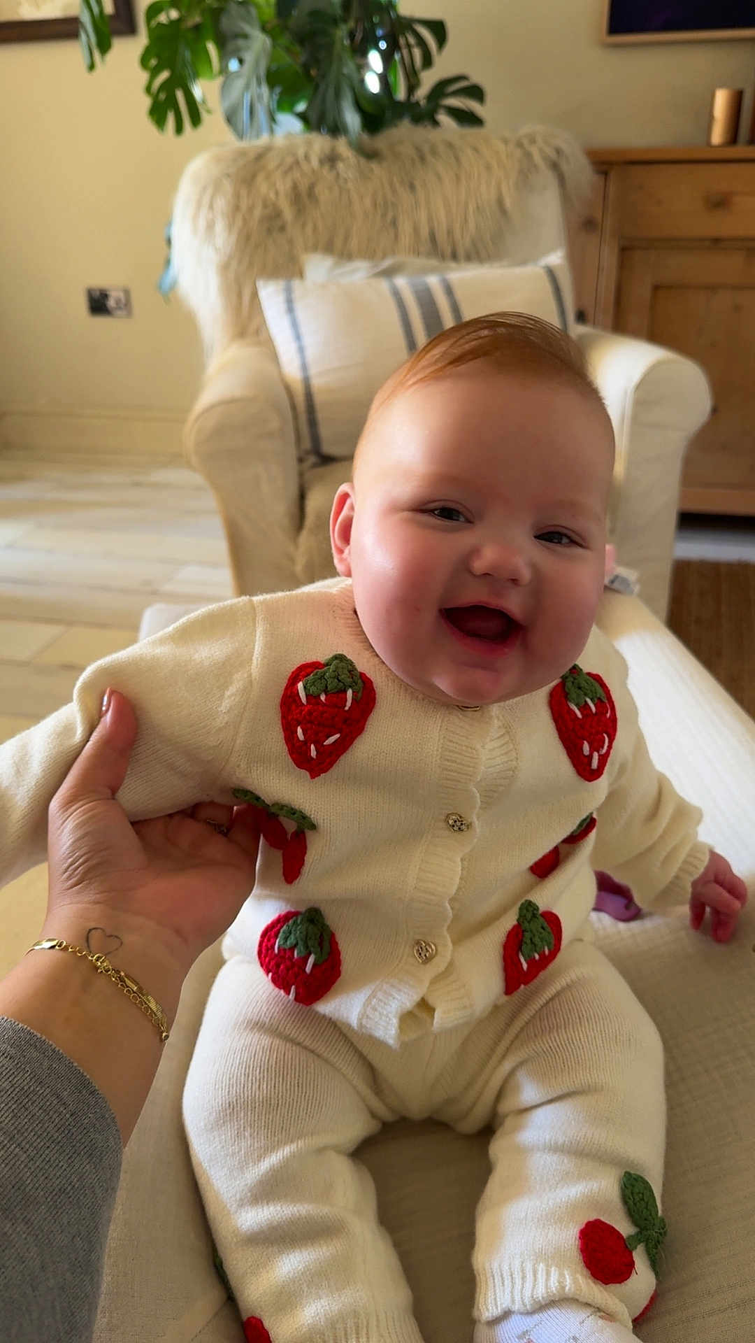 Matilda is registered to the contest to win money with this photo: baby, child, smiling, laughing, person, indoor, couch, armchair, hand, tattoo, bracelet, knitwear, strawberry_pattern, cozy, home, furniture, pillow, plant, wooden_furniture, floor