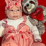 baby, infant, pink_dress, headband, bow, plush_toy, sloth, heart_pattern, valentines_theme, red_background, cute, soft_toy, child, smiling, lying_down, toddler, clothing, love, stuffed_animal, portrait