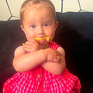 Ariella is registered to the contest to win money with this photo: baby, child, infant, dress, red, checkered, couch, snack, food, hair, ponytail, sitting, indoor, face, hands, cute, curious, patterned_wall, person, young