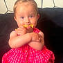 baby, child, infant, dress, red, checkered, couch, snack, food, hair, ponytail, sitting, indoor, face, hands, cute, curious, patterned_wall, person, young