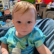 Leon is registered to the contest to win money with this photo: baby, child, bottle, tie_dye_shirt, indoor, person, face, eyes, hair, hand, clothing, furniture, couch, carpet, living_room, baby_bottle, holding, sitting, curious, young