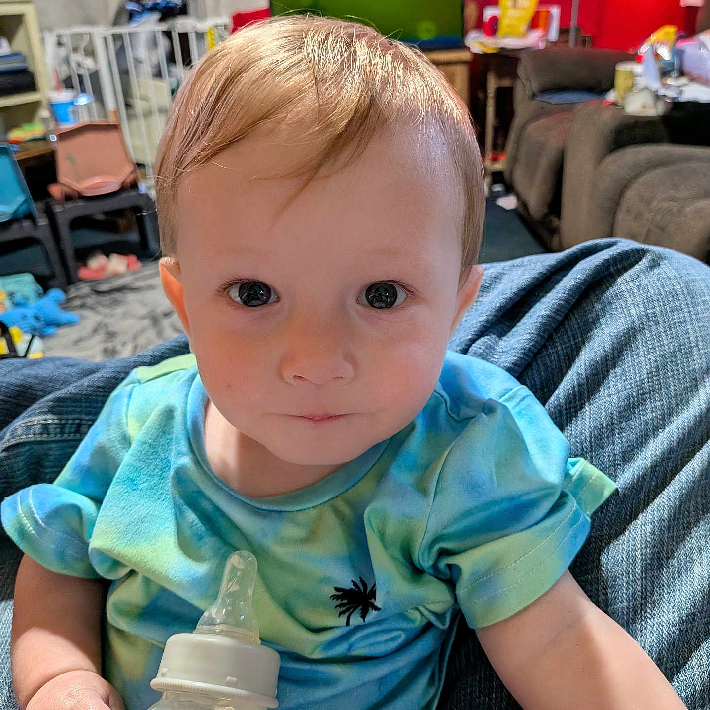 Leon is registered to the contest to win money with this photo: baby, child, bottle, tie_dye_shirt, indoor, person, face, eyes, hair, hand, clothing, furniture, couch, carpet, living_room, baby_bottle, holding, sitting, curious, young