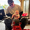 baby, cabinet, cap, chair, clothing, face, garage, handlebars, hands, indoor, jeans, light, motorbike, person, shirt, sticker, sunglasses, toy_vehicle, window, young_adult