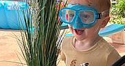 Coltyn is registered to the contest to win money with this photo: child, toddler, goggles, plant, potted_plant, outdoor, patio, pool, umbrella, fence, wooden_post, grass, sunlight, smiling, clothing, short_sleeve_shirt, toy, happy, summer, backyard