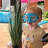 child, toddler, goggles, plant, potted_plant, outdoor, patio, pool, umbrella, fence, wooden_post, grass, sunlight, smiling, clothing, short_sleeve_shirt, toy, happy, summer, backyard