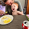toddler, child, adult, feeding, scrambled_eggs, banana, plate, table, smile, laughing, red_hair, smartphone, can, coca_cola, indoor, casual, breakfast, family, happy, person