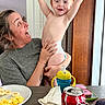 child, toddler, woman, diaper, breakfast, scrambledeggs, banana, sippy_cup, coca_cola_can, smartphone, table, happy, smile, indoor, person, food, napkin, glasses, holding, joyful