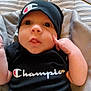 baby, infant, clothing, hat, black_shirt, cushion, seat, face, expression, hand, person, portrait, indoors, cute, child, sitting, apparel, logo, human, babywear