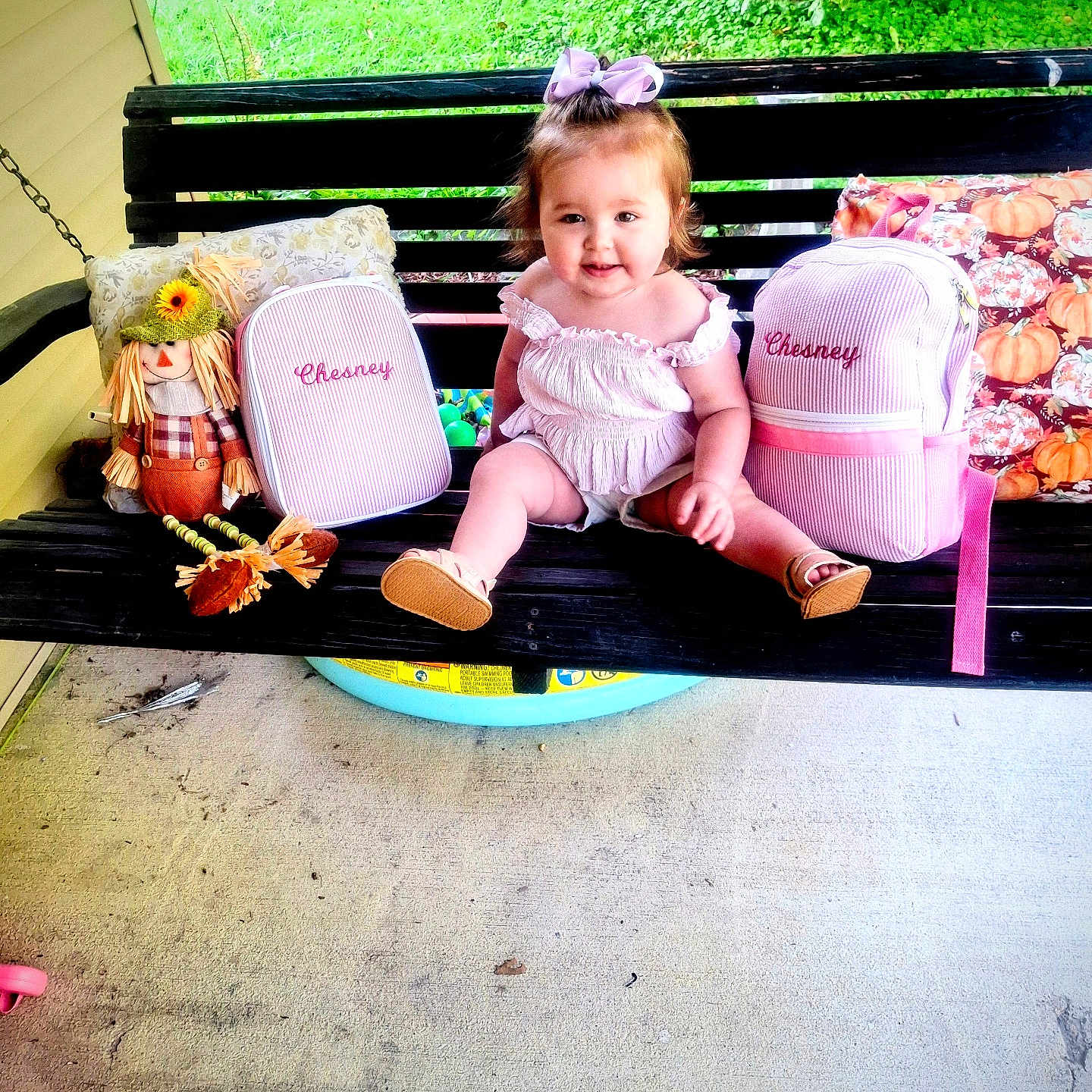 Chesney joined the competition — help win amazing prizes! black_swing, child, concrete_floor, cute, decorative_pillows, embroidered_name, green_grass, happy, outdoor, pink_bags, playful, porch_swing, purple_bow, scarecrow_doll, sitting, smiling, summer, sunlight, toddler, white_outfit