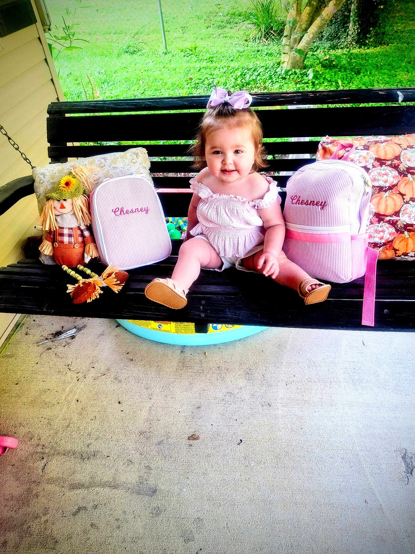 Chesney joined the competition — help win amazing prizes! toddler, child, porch_swing, black_swing, pink_bags, embroidered_name, scarecrow_doll, decorative_pillows, green_grass, outdoor, concrete_floor, purple_bow, white_outfit, smiling, sitting, summer, sunlight, cute, happy, playful