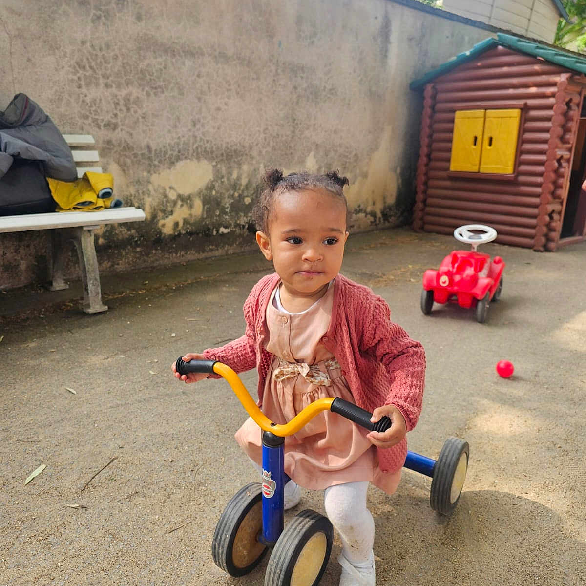 Rose-Esther a rejoint le concours — aidez-le/la à gagner de superbes lots ! bench, cardigan, child, curly_hair, daylight, jacket, outdoor, pigtails, pink_dress, play, playground, playhouse, ride_on_car, sand, toddler, toy, tricycle, white_shoes, white_tights, young_child