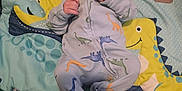 Hakeem is registered to the contest to win money with this photo: baby, dinosaur, onesie, blanket, infant, cute, child, lying_down, pattern, colorful, soft, playful, indoor, young, person, smiling, hand, feet, fabric, bedtime