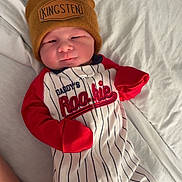 Kingsten joined the competition — help win amazing prizes! baby, infant, beanie, hat, onesie, striped_clothing, red_sleeves, bed, blanket, footwear, socks, person, smiling, sleepwear, cute, indoor, relaxing, child, young, portrait