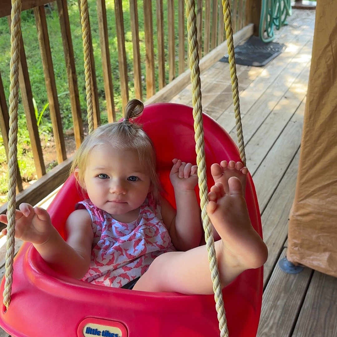 Koura is registered to the contest to win money with this photo: toddler, child, swing, red_swing, porch, wooden_floor, outdoor, rope, greenery, sunlight, playful, barefoot, topknot, summer, daylight, cute, person, smiling, relaxed, home