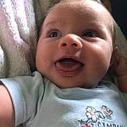Oakley Jade is registered to the contest to win money with this photo: baby, infant, smiling, tongue_out, blue_shirt, text_on_clothing, blanket, close_up, face, cute, happy, child, soft_texture, lying_down, portrait, skin, young_child, indoors, expression, person