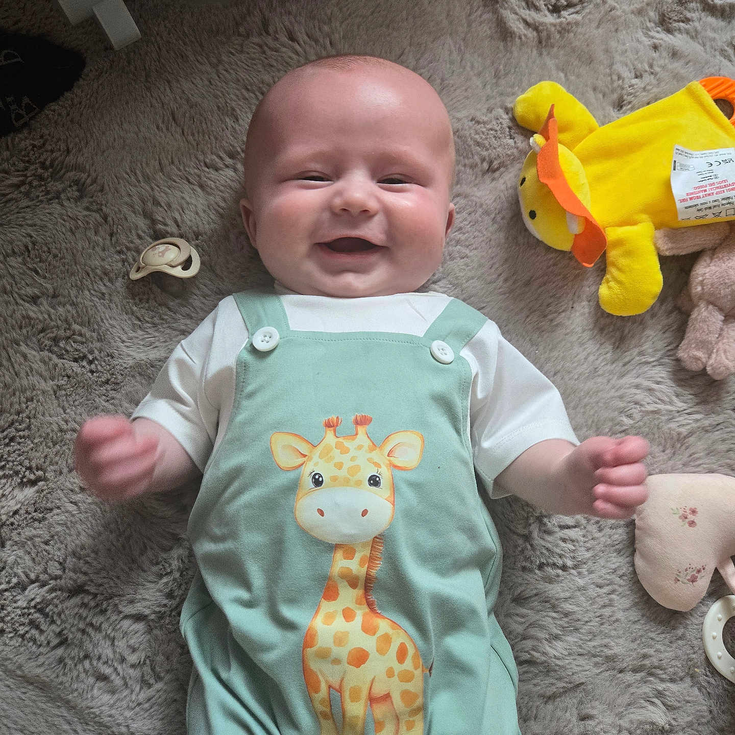 Jaxx joined the competition — help win amazing prizes! baby, infant, smiling, giraffe_print, plush_toy, pacifier, soft_rug, green_outfit, white_shirt, cute, happy, indoors, child, toy, floor, playtime, cozy, comfort, lying_down, portrait