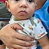 baby, infant, child, portrait, wide_eyes, serious_expression, sitting, being_held, adult, caregiver, onesie, patterned_clothing, trucks, trees, indoor, close_up, face, hands, family, photography