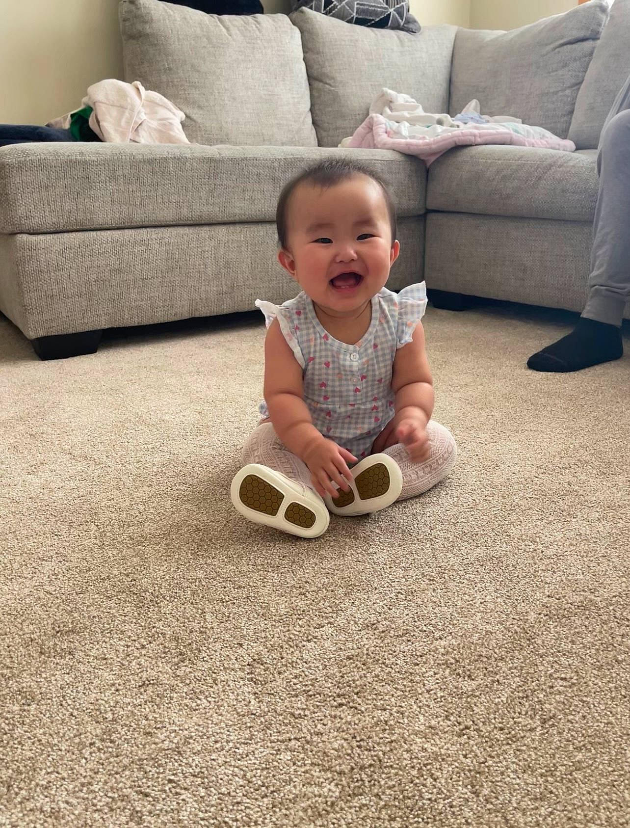 Seraphine is registered to the contest to win money with this photo: baby, baby_toddler_clothing, comfort, couch, floor, flooring, happy, hardwood, human_leg, knee, leg, living_room, person, pillow, product, sleeve, smile, textile, toddler, white