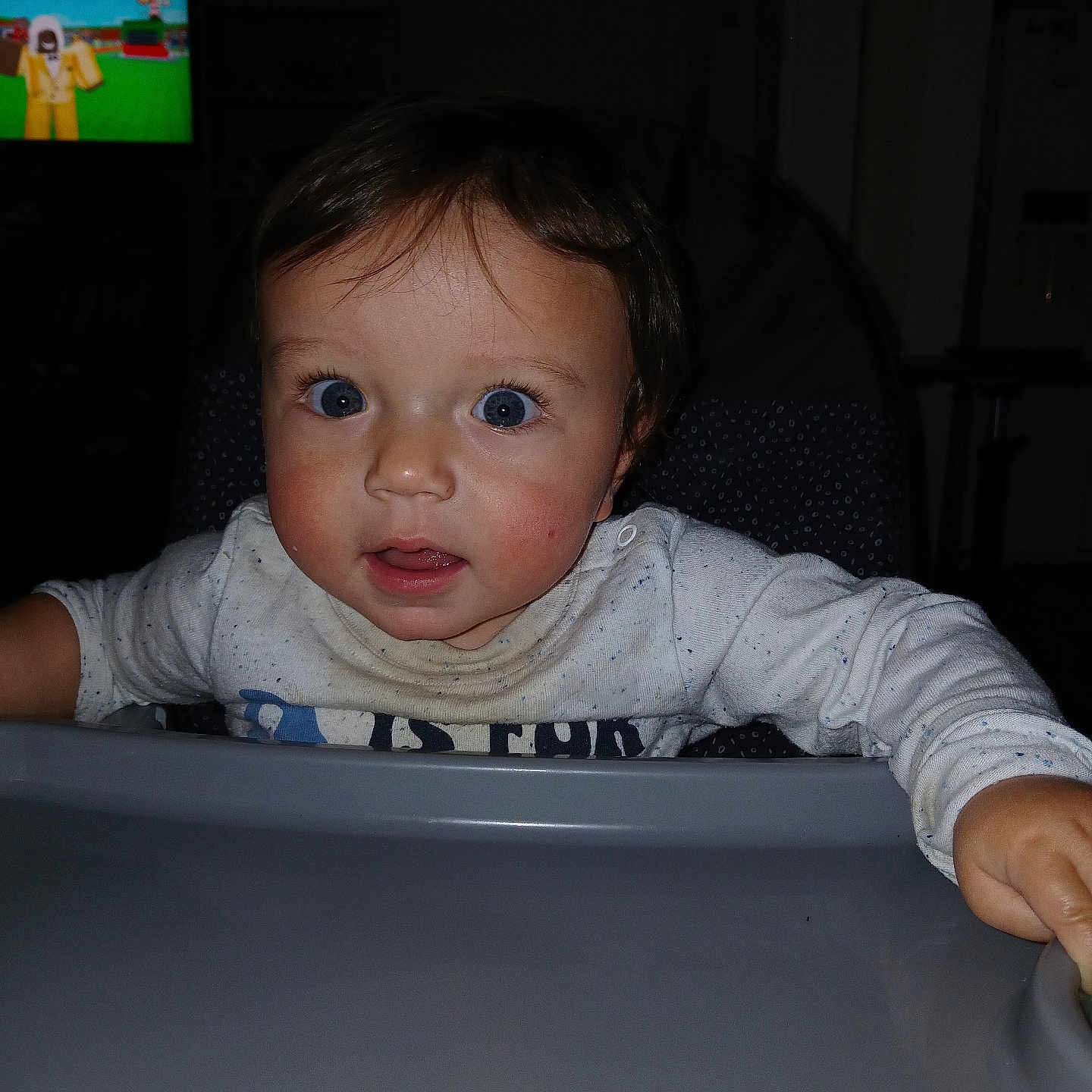 Jeremiah is registered to the contest to win money with this photo: baby, infant, child, high_chair, portrait, face, eyes, wide_eyed, sitting, indoor, home, dining_area, tray, shirt, sleeves, television, cartoon, dark_room, flash_photography, close_up