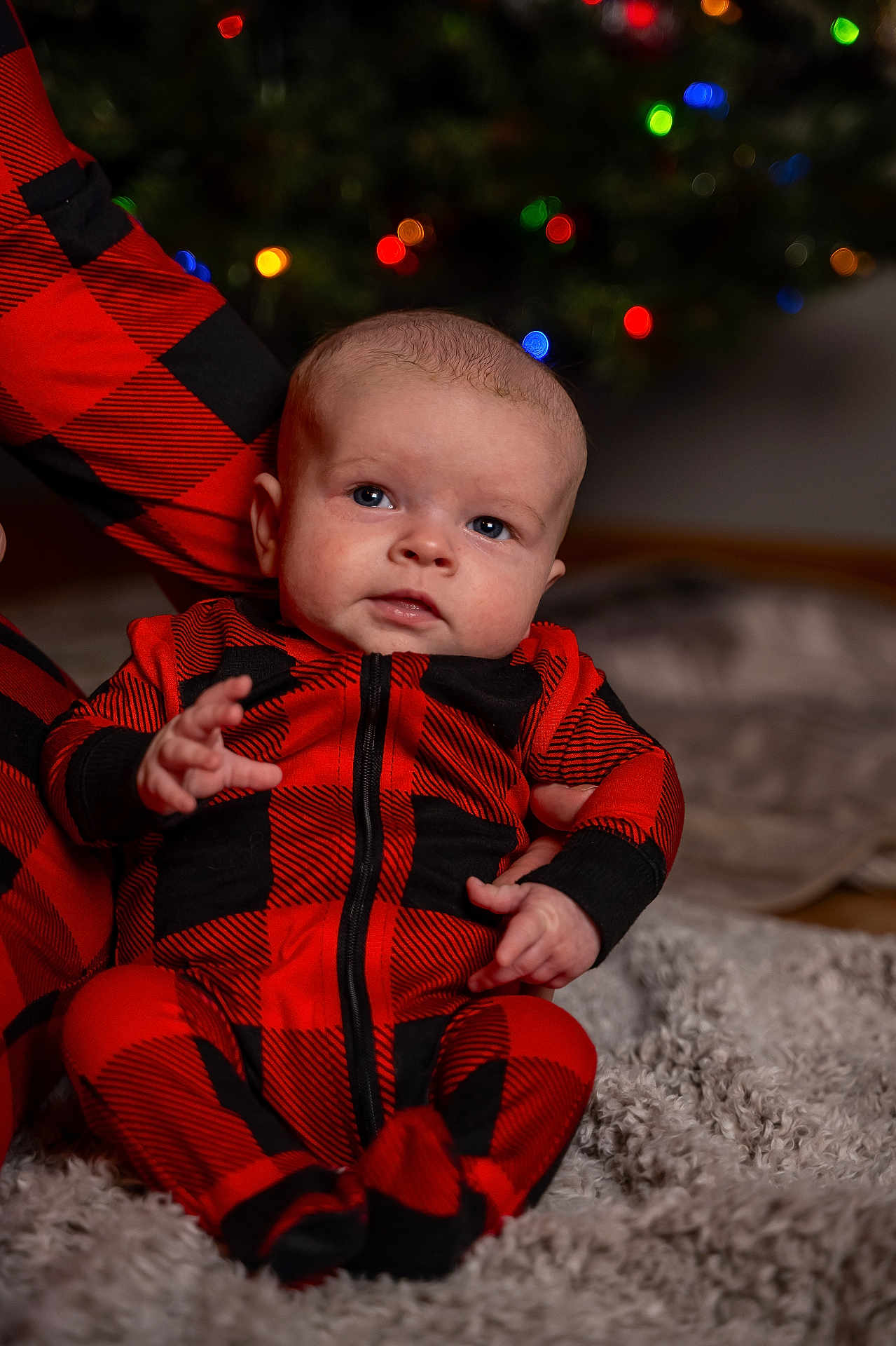 Liam is registered to the contest to win money with this photo: baby, infant, red_clothing, black_clothing, plaid_pattern, onesie, blanket, holiday_lights, christmas, blurred_background, cute, child, indoor, portrait, sitting, hand, face, head, soft_texture, cozy
