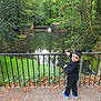 child, boy, outdoor, nature, pond, trees, greenery, railings, path, leaves, fall, water, reflection, casual_clothing, sneakers, curious, standing, daylight, forest, scenery
