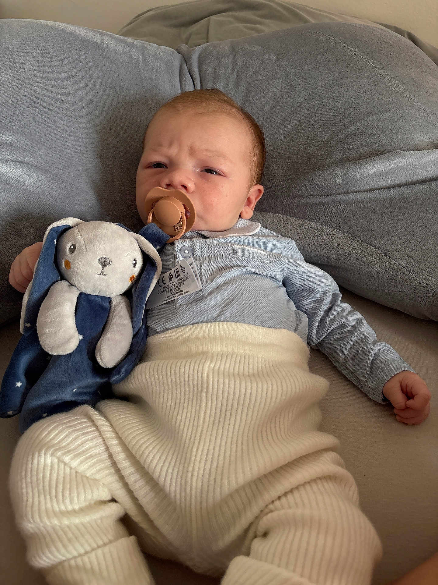 Sohan a rejoint le concours — aidez-le/la à gagner de superbes lots ! baby, pacifier, plush_toy, bunny, blue_clothing, cream_pants, cushion, soft_texture, indoor, infant, child, cute, lying_down, serious_expression, toy, comfort, cozy, face, person, home