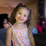 Ariella is registered to the contest to win money with this photo: child, girl, smiling, curly_hair, pink_bows, dress, colorful, indoors, living_room, toys, furniture, carpet, person, cute, happy, portrait, young_child, playful, home, casual_clothing