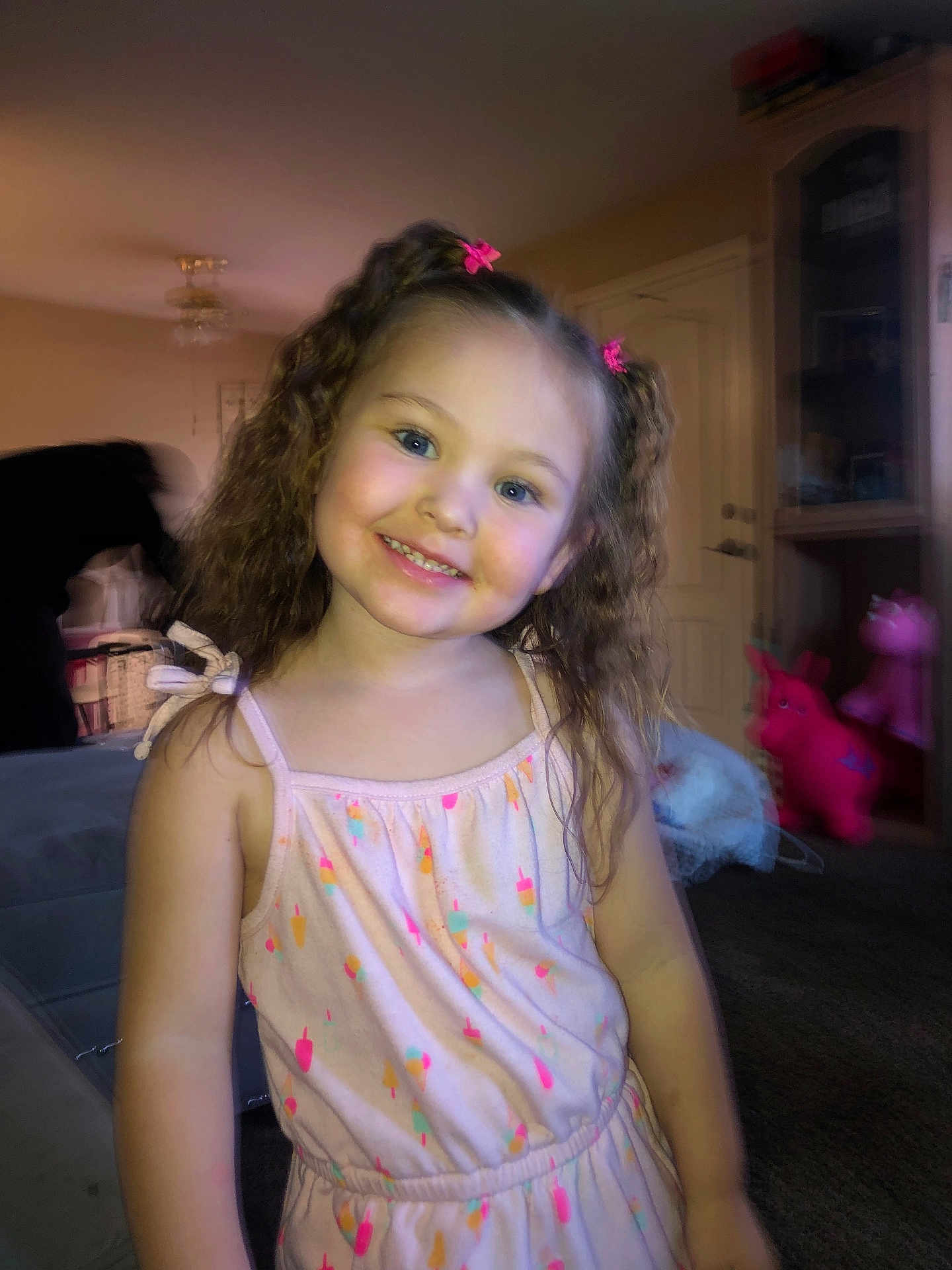 Ariella is registered to the contest to win money with this photo: child, girl, smiling, curly_hair, pink_bows, dress, colorful, indoors, living_room, toys, furniture, carpet, person, cute, happy, portrait, young_child, playful, home, casual_clothing