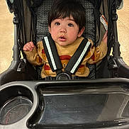 Rameses is registered to the contest to win money with this photo: toddler, child, stroller, sweater, bear_pattern, curious_expression, seatbelt, indoor, tray, cup_holder, black_hair, sitting, baby, clothing, person, young_child, face, looking_up, cozy, casual