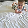 baby, infant, bed, white_bed_sheet, lying_down, curious, indoor, headboard, nightstand, sleepwear, shorts, patterned_shirt, skin, face, eyes, smile, hand, foot, furniture, room