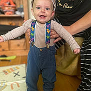 Ernie joined the competition — help win amazing prizes! toddler, child, smiling, cap, suspenders, dinosaur_pattern, striped_shirt, blue_pants, socks, indoor, wooden_floor, furniture, adult_hands, supporting, happy, person, playful, expression, standing, home