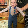 toddler, child, smiling, cap, suspenders, dinosaur_pattern, striped_shirt, blue_pants, socks, indoor, wooden_floor, furniture, adult_hands, supporting, happy, person, playful, expression, standing, home
