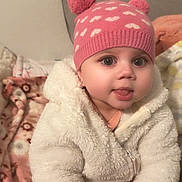 Gianna joined the competition — help win amazing prizes! baby, child, pink_hat, pom_poms, white_jacket, fluffy_clothing, blanket, patterned_blanket, indoor, cute, tongue_out, smiling, warm_clothing, seated, infant, face, hands, eyes, cozy, young_child