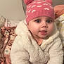 baby, child, pink_hat, pom_poms, white_jacket, fluffy_clothing, blanket, patterned_blanket, indoor, cute, tongue_out, smiling, warm_clothing, seated, infant, face, hands, eyes, cozy, young_child